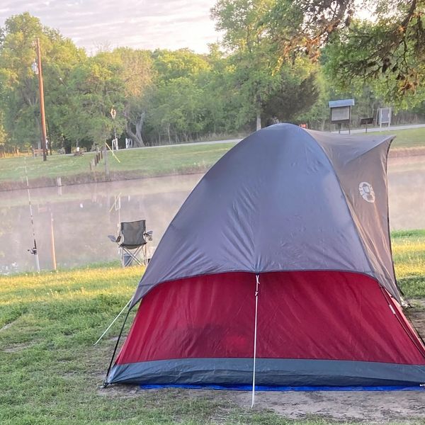 A red and gray tent set up near a calm lake with a chair and fishing rods nearby.