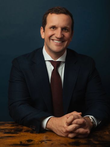 A smiling man in a dark suit and red tie sitting at a wooden table.