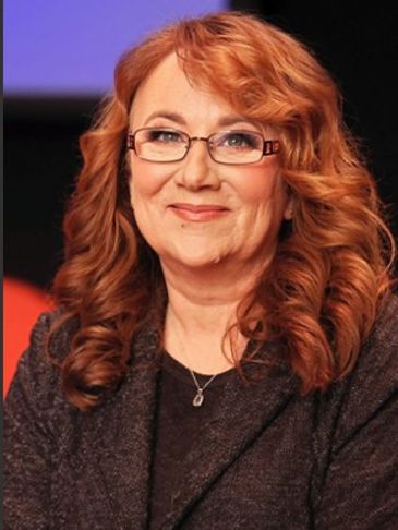 Smiling woman with curly red hair and glasses in a dark outfit.