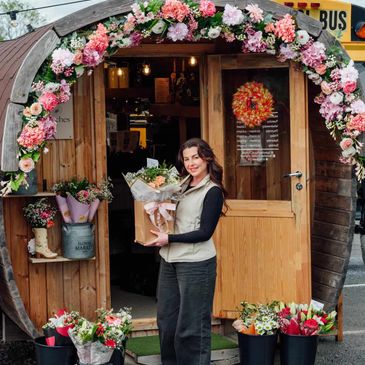Woman holding a bouquet standing in front of a flower shop with floral arch.