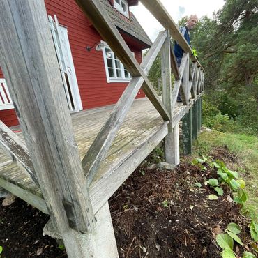 Man standing on the wooden deck of a red house surrounded by greenery.