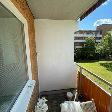 Cozy balcony with wooden flooring, a chair, flowers, and a sunny view of green grass and buildings.
