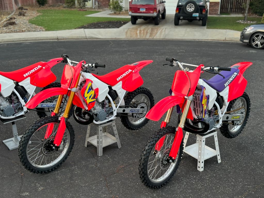 Two red Honda dirt bikes on stands in a driveway.