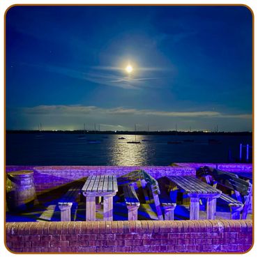 Moonlit waterfront with wooden benches and tables illuminated in blue light.