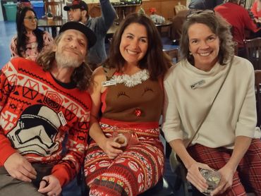 Three friends smiling and sitting together in festive holiday attire indoors.