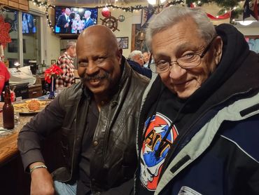 Two elderly men smiling together at a cozy bar with holiday decorations.
