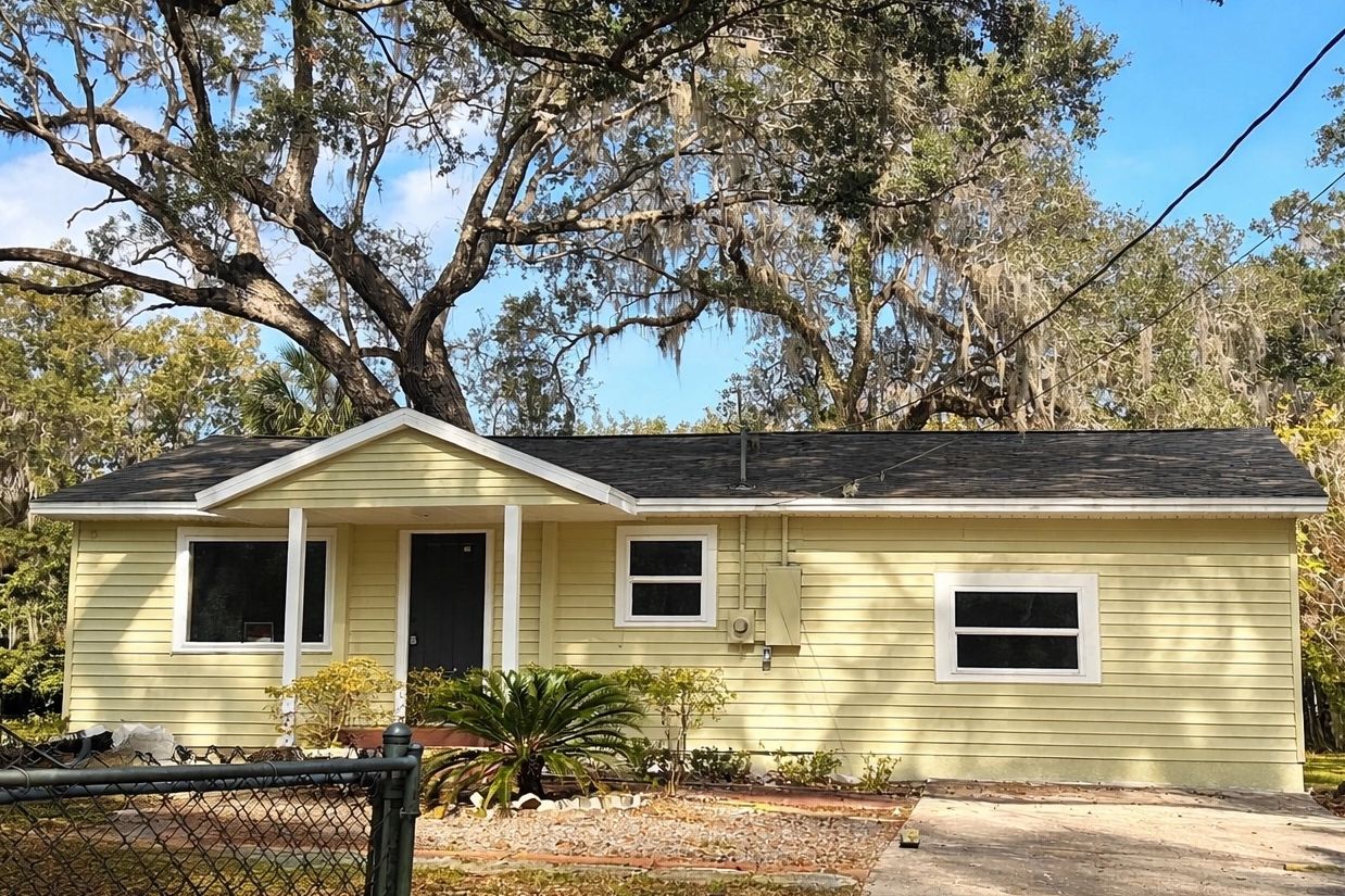 Small yellow house with a front porch and large trees.
