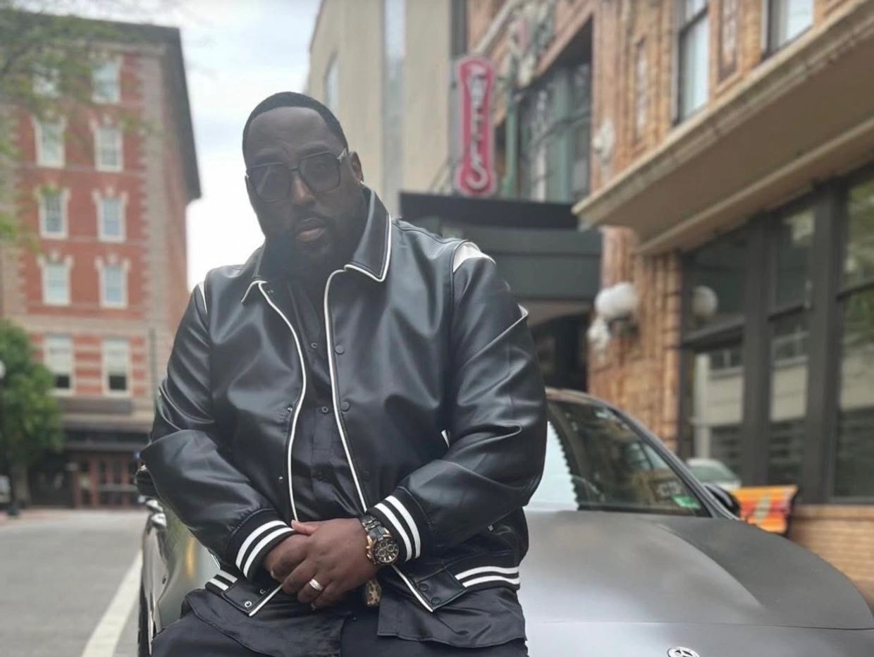 Man in black leather jacket sitting on a car in an urban setting.