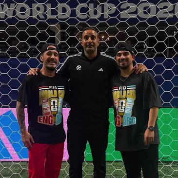 Three men smiling in front of a soccer goal with FIFA World Cup shirts.