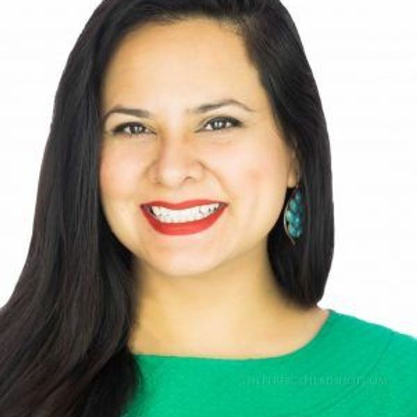 Smiling woman with long dark hair and red lipstick, wearing green top and turquoise earrings.