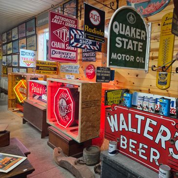A collection of vintage advertising signs displayed on a wooden wall. The neon signs represent Walter's Beer, Oshkosh, Point Beer, and Grainbelt. There are also gas and oil signs and thermometers.