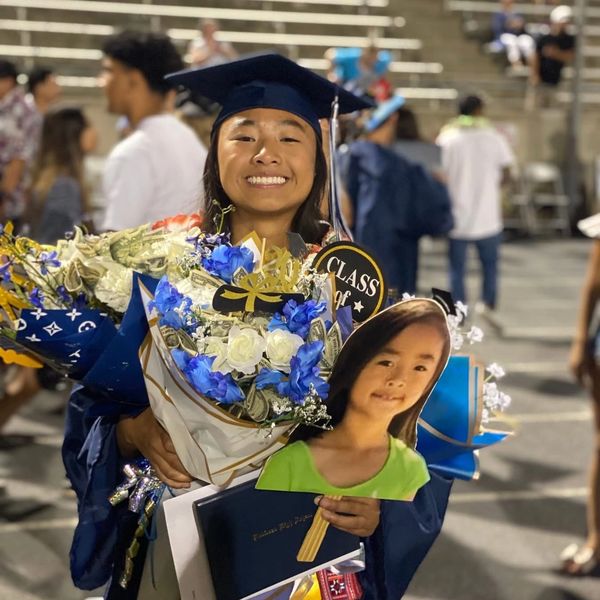 Smiling graduate in cap and gown holding flowers and a childhood photo cutout.