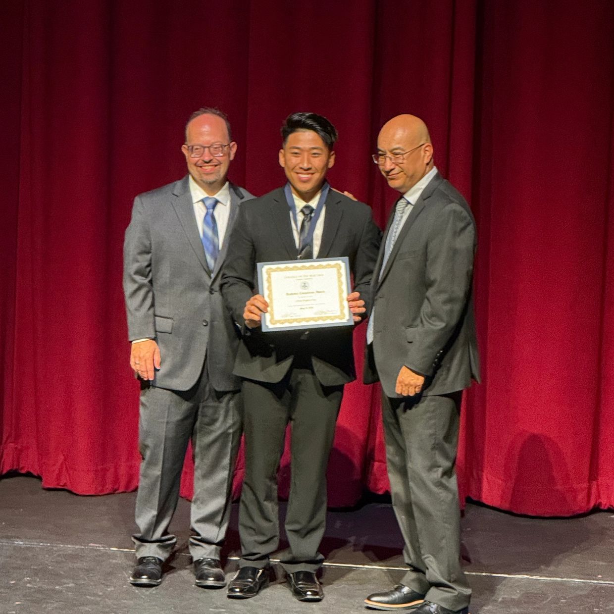Three men in suits, one holding an award certificate on stage with red curtains.