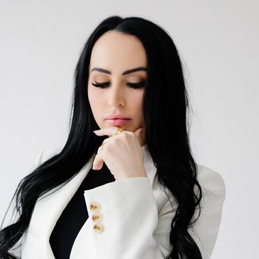 Woman with long black hair in white blazer, looking thoughtful.
