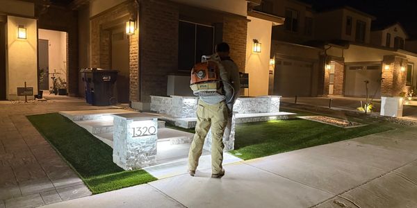A person with a leaf blower stands outside a lit modern home at night.