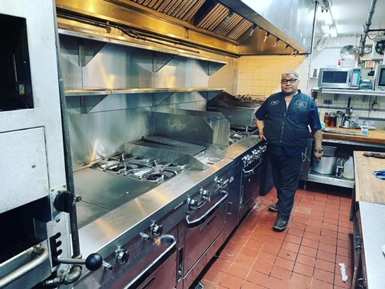 Man standing in a large commercial kitchen beside stainless steel cooking equipment.