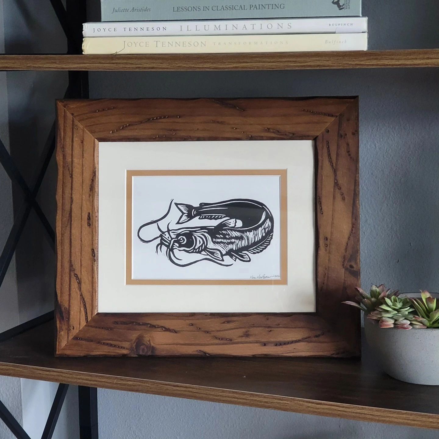 Framed black and white fish illustration on a wooden shelf with books and a succulent.