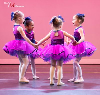 Four young girls in purple tutus holding hands in a dance circle on stage.