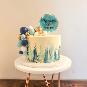 Single tier cake with blue strokes detail, ball decorations, gold sprinkles and cake topper.