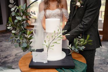 Close up of couple cutting 3 tier white wedding cake with foliage detail