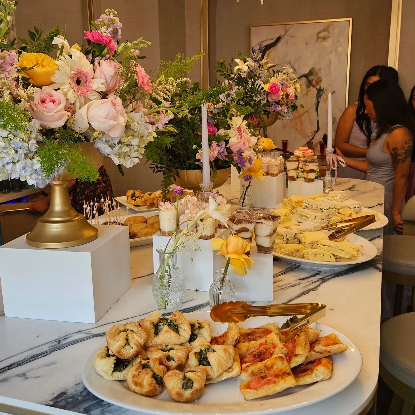 Elegant buffet table with pastries, sandwiches, desserts, and floral arrangements.