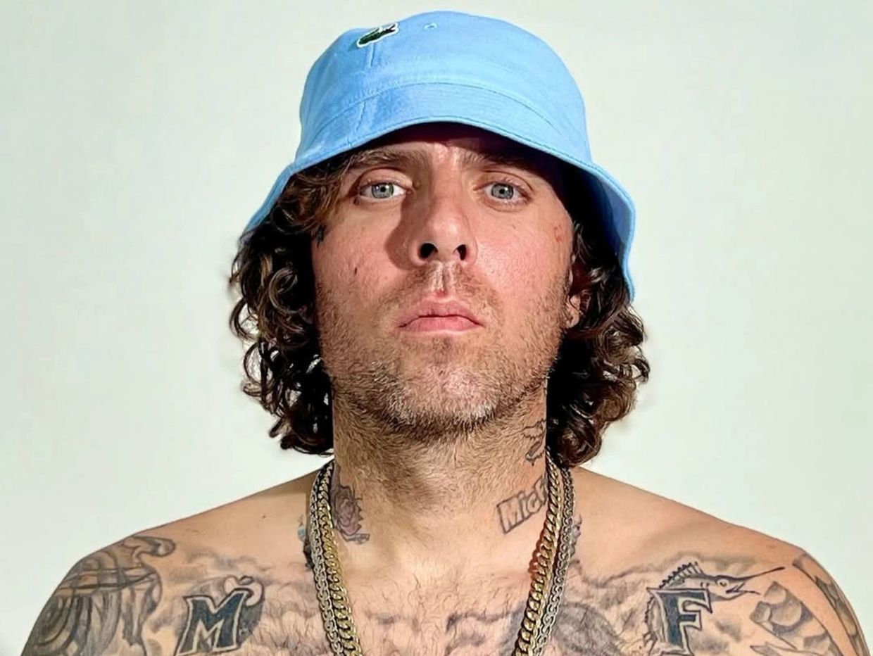 Man with curly hair and tattoos wearing a blue bucket hat and gold chains.