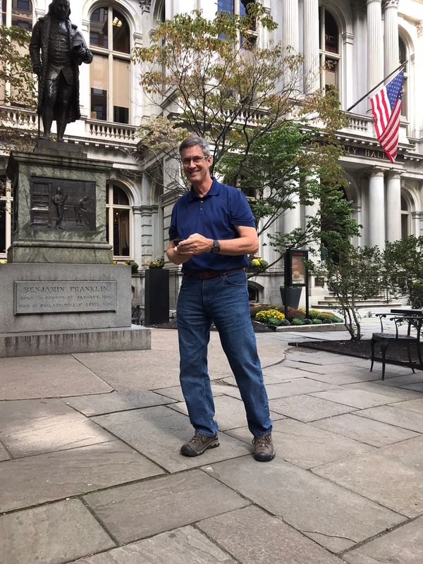 Man in blue shirt stands near Benjamin Franklin statue outside city hall.