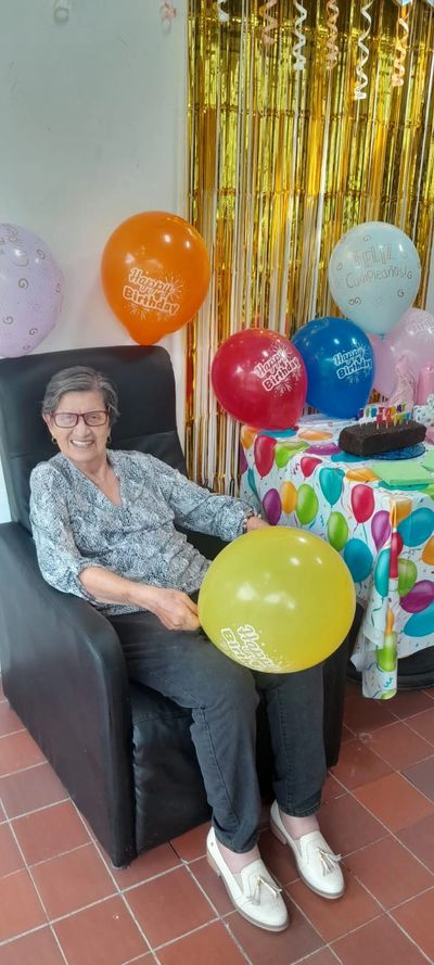 Elderly woman smiling while holding a yellow birthday balloon, sitting near a decorated table with colorful balloons and cake.