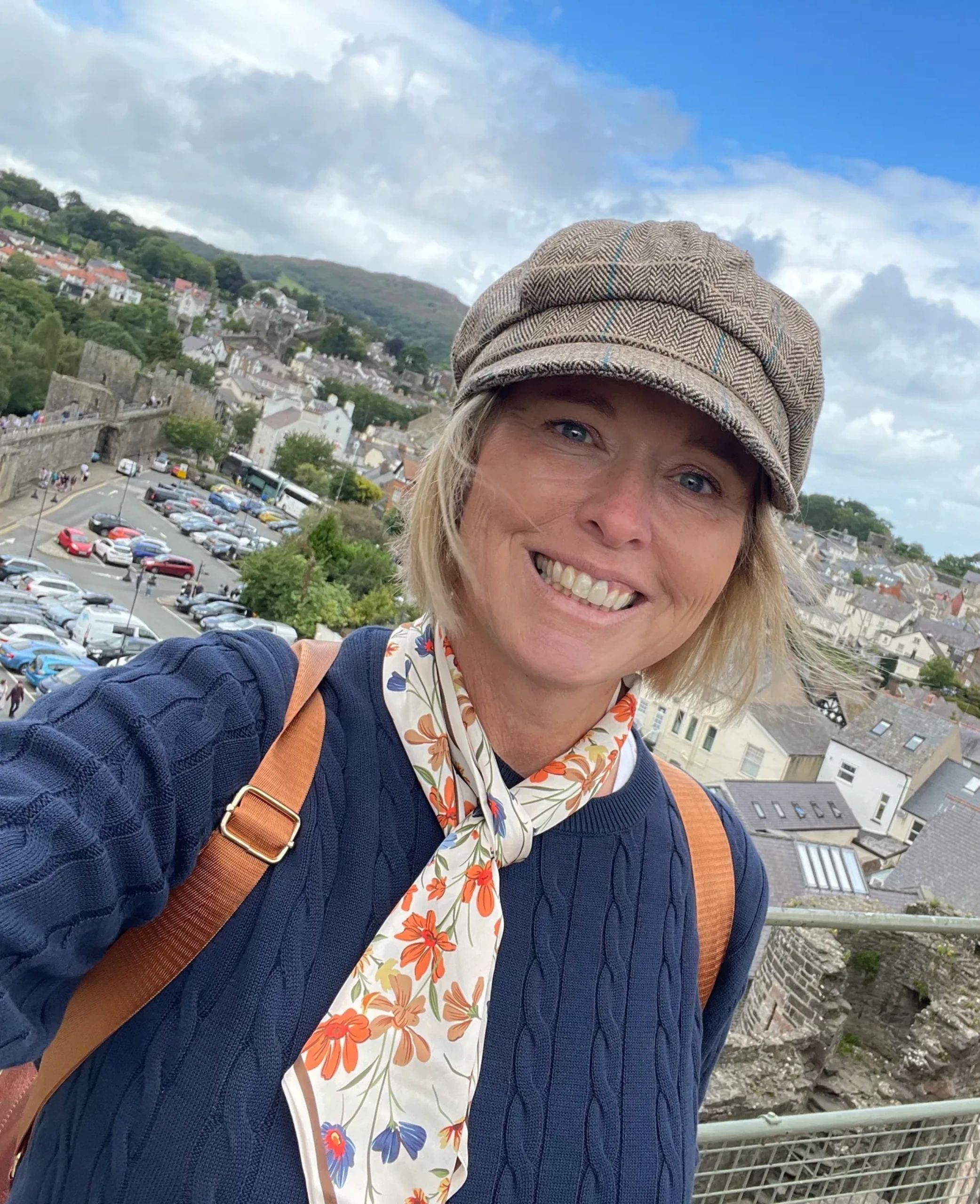 Smiling woman wearing a patterned cap and floral scarf with a scenic town in the background.