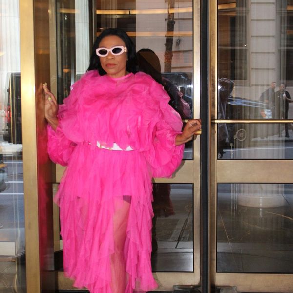 Woman in a vibrant pink ruffled dress and white sunglasses exiting a building.