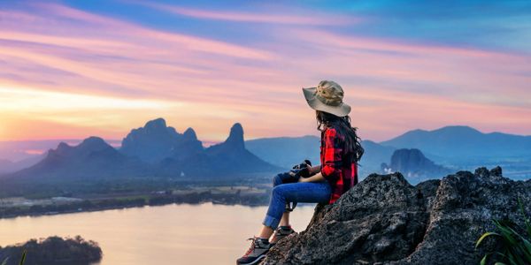 Woman in hat sitting on rock, enjoying a colorful sunset over mountains and a river.