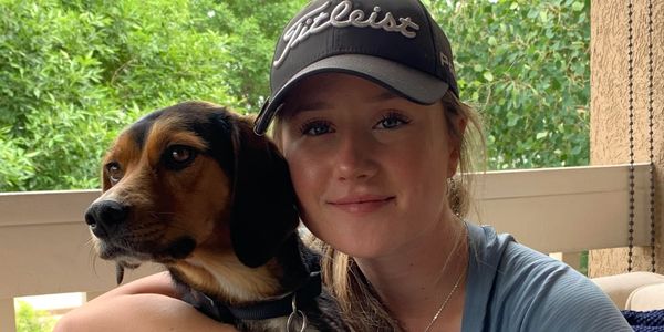 Young woman in a cap hugging a dog on a porch with greenery behind.