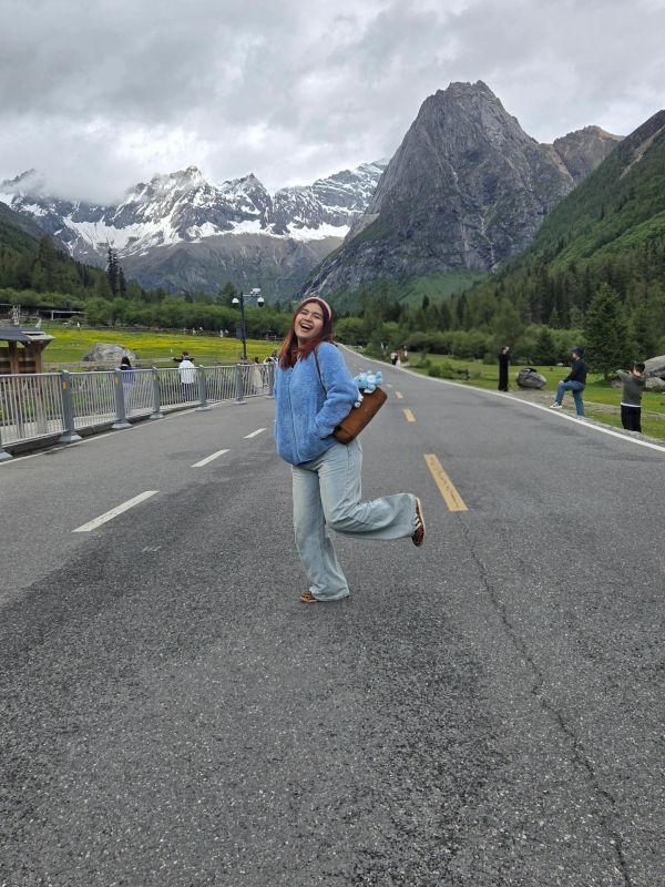 A joyful woman poses on a mountain road with snowy peaks in the background.