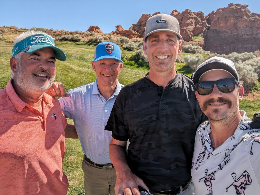 Four men smiling on a sunny golf course with rocky hills in the background.