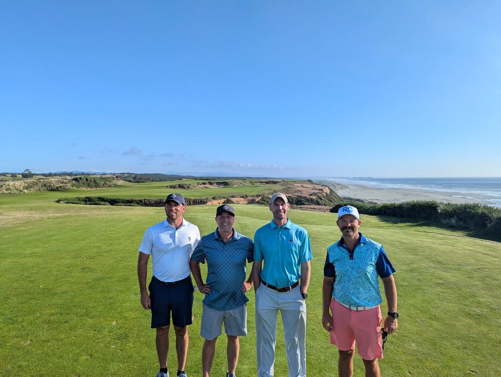 Four men standing on a golf course with ocean and blue sky in the background.