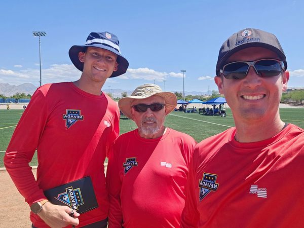 Three men wearing red shirts and hats posing on a sports field under clear blue sky.