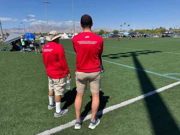 Two people in red shirts watch a sports game on a sunny field.
