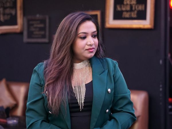 Woman in a green blazer standing indoors with folded hands.