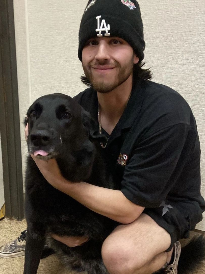 Young man in LA beanie hugging a black dog indoors.