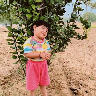 A smiling child stands by a leafy tree in a rural outdoor setting.