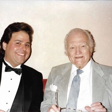 Two men smiling, one in a tuxedo and the other in a grey suit.
