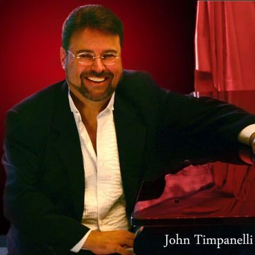 Smiling man in a black suit seated at a piano with red backdrop.