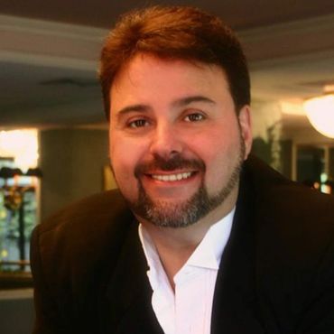 Smiling man with beard in a formal black jacket indoors.