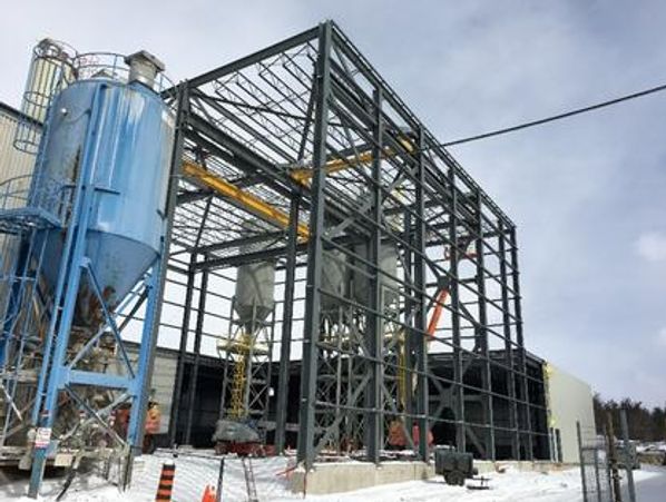 Construction of an industrial factory building with exposed steel framework and a blue silo.
