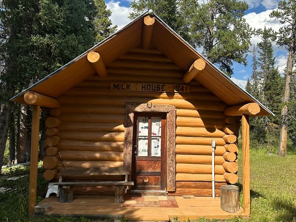 Rustic log cabin labeled 'Milk House 1941' in a forest clearing.