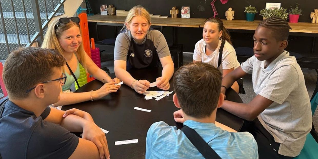 A group of six teenagers sitting around a table, interacting and holding paper slips.