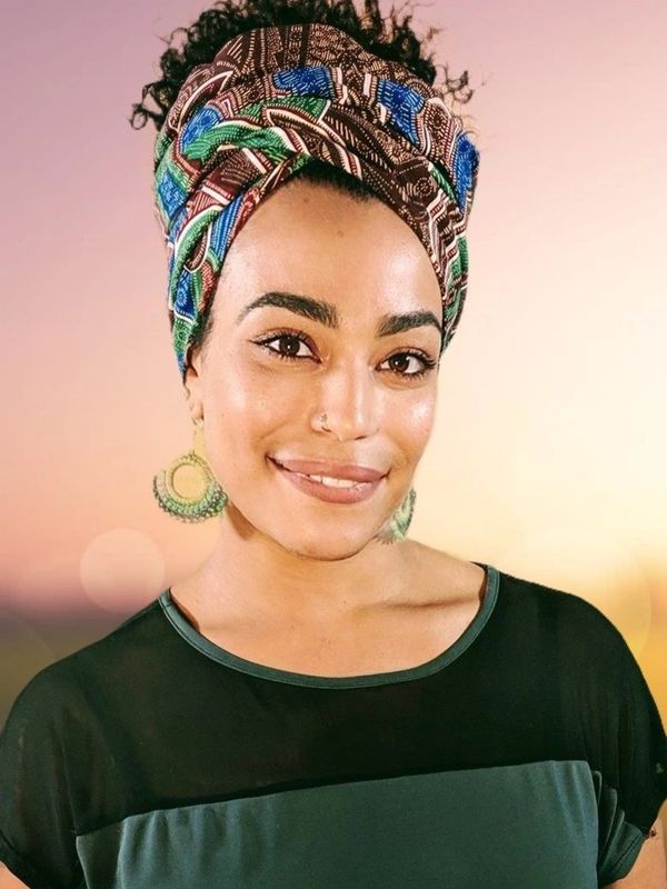 Smiling woman with colorful headwrap and large earrings against a soft sunset background.
