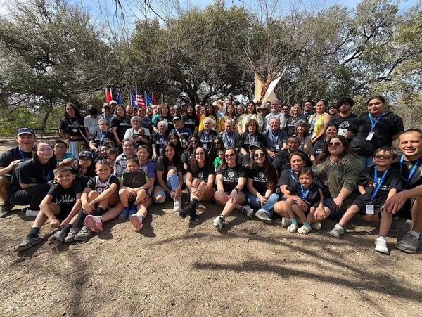Large diverse group posing outdoors under trees with flags and teepees in background.