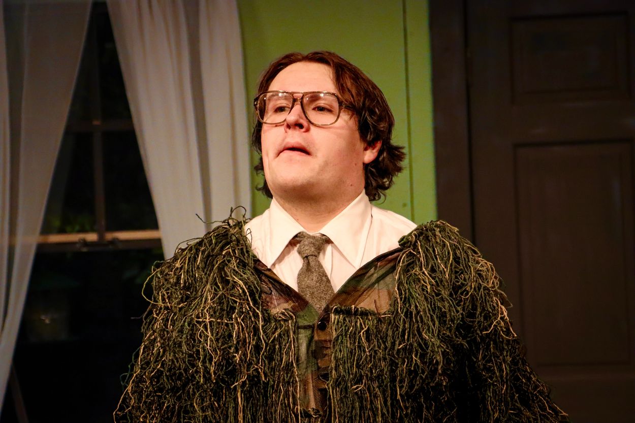 A man in glasses wearing a leafy camouflage jacket over formal attire indoors.