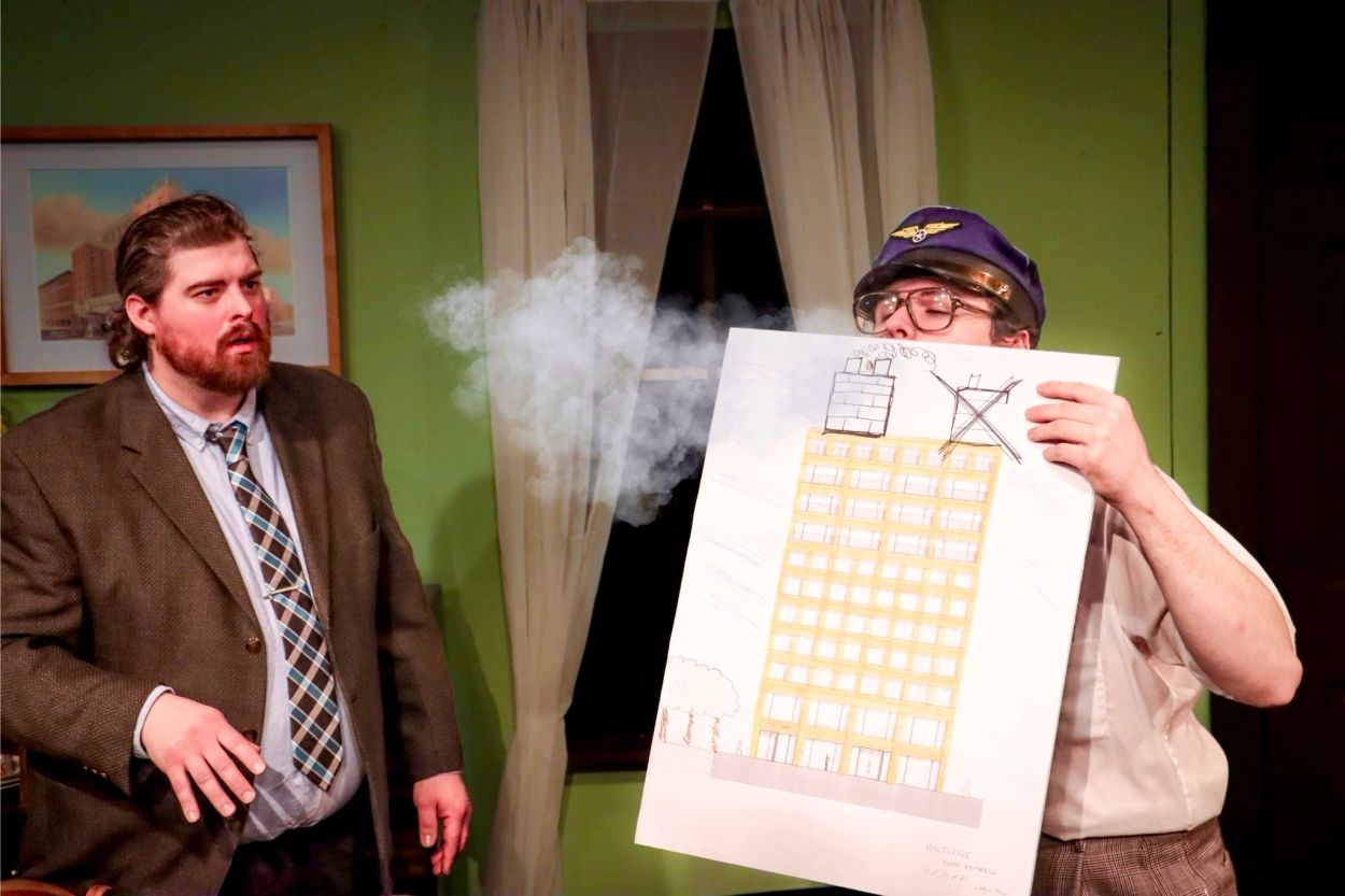 Two men perform a comedic scene with a smoke effect and a building drawing.